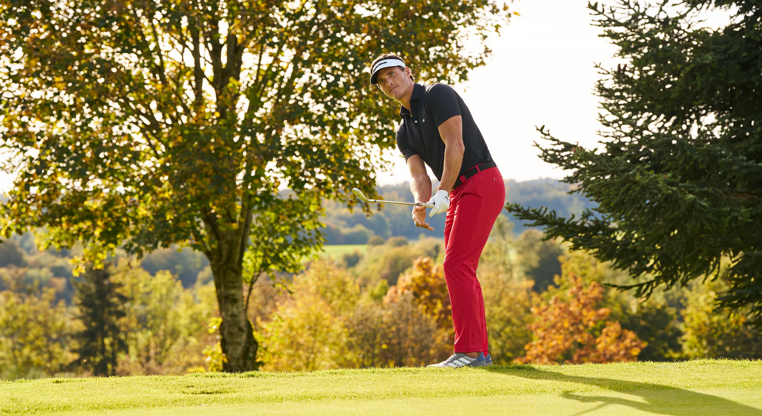 Golfer in red pants and black shirt swinging a golf club on a golf course with trees in the background.